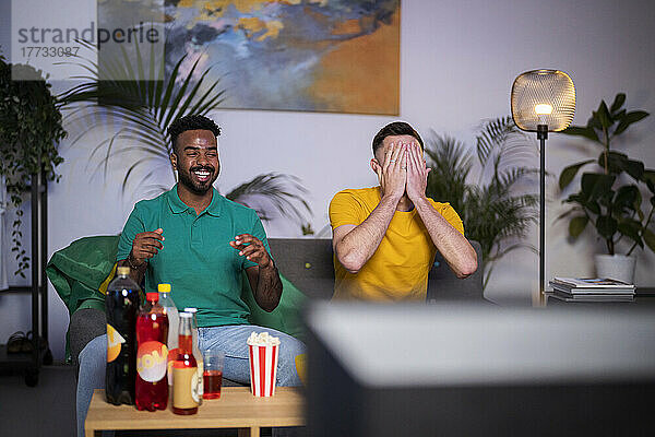 Sad man covering face sitting by happy friend watching football match at home