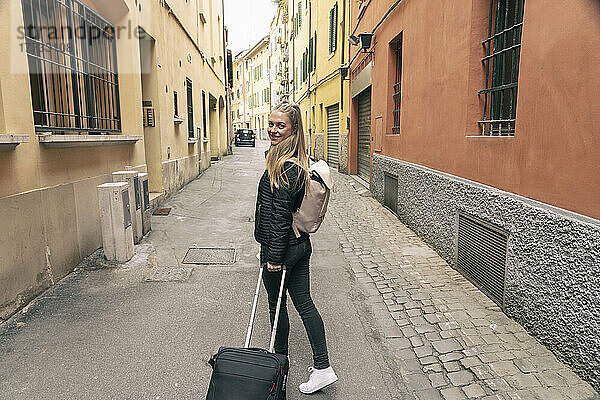 Lächelnde blonde Frau mit Rucksack läuft mit Rollgepäck durch Gebäude