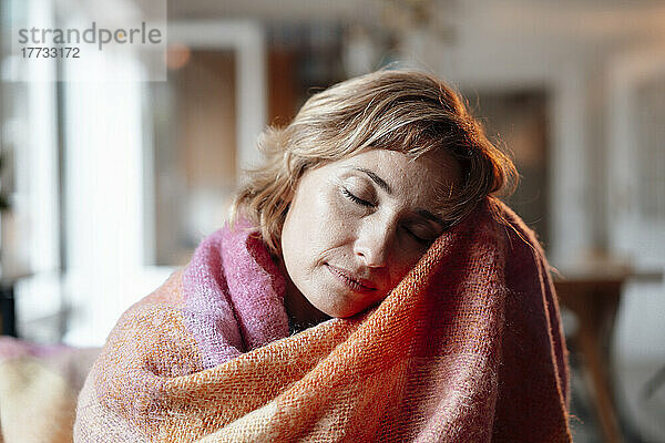 In eine Decke gehüllte Frau sitzt mit geschlossenen Augen zu Hause