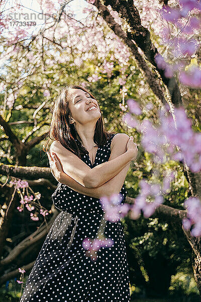 Lächelnde junge Frau mit geschlossenen Augen umarmt sich im Garten