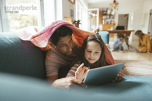 Mädchen benutzt Tablet-PC und liegt mit Vater zu Hause auf dem Sofa