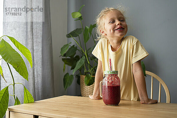 Glückliches Mädchen  das zu Hause beim Smoothie auf dem Tisch steht