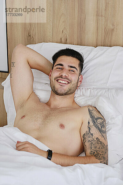 Happy shirtless man with hand behind head in bed at home