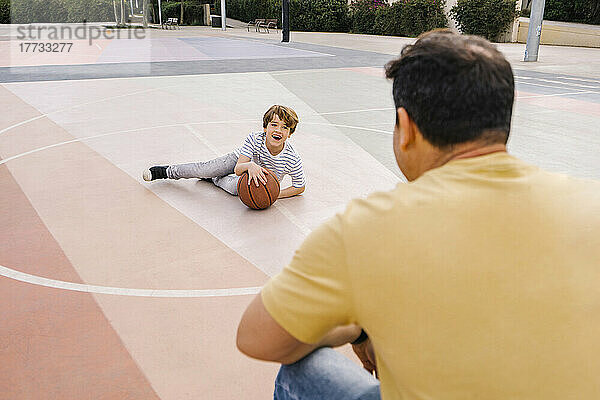 Glücklicher Junge mit Basketball sitzt vor seinem Vater auf dem Sportplatz