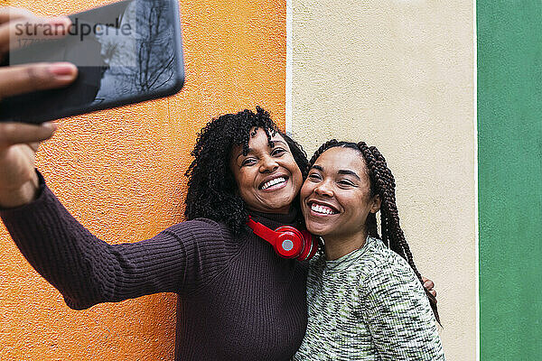 Fröhliche Frau mit Freundin macht Selfie mit Smartphone vor der Wand