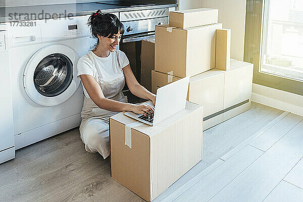 Glückliche Frau benutzt Laptop auf Karton neben Waschmaschine im Hauswirtschaftsraum