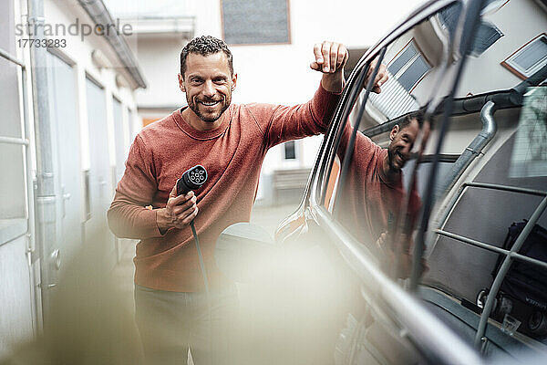Lächelnder Mann hält Ladekabel in der Hand und stützt sich auf ein Elektroauto