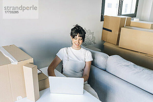 Lächelnde Frau mit Laptop sitzt mit Kartons am Tisch im Wohnzimmer