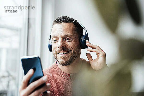 Lächelnder Mann mit Kopfhörern  der zu Hause sein Smartphone benutzt
