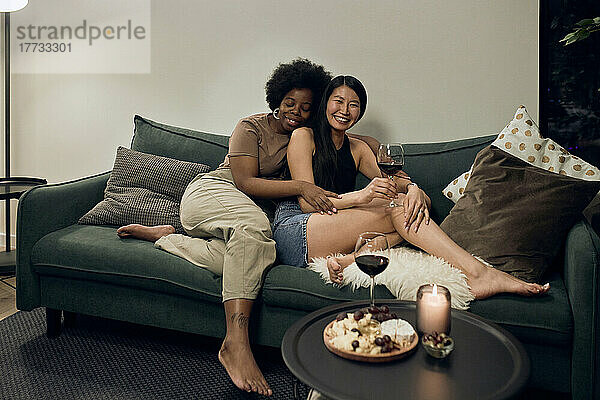 Lächelnde junge Frau umarmt ihre Freundin  die zu Hause mit Weinglas auf dem Sofa sitzt