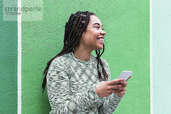 Glückliche junge Frau mit Smartphone steht vor grüner Wand