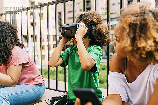 Junge mit Afro-Frisur trägt einen Virtual-Reality-Simulator und sitzt inmitten von Freunden