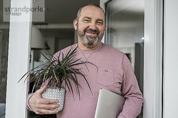 Lächelnder Geschäftsmann mit Drachenbaum und Laptop vor der Tür