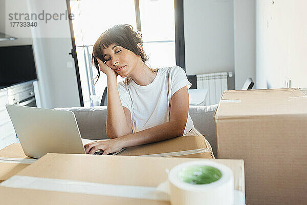 Müde Frau mit Laptop auf Karton im neuen Zuhause