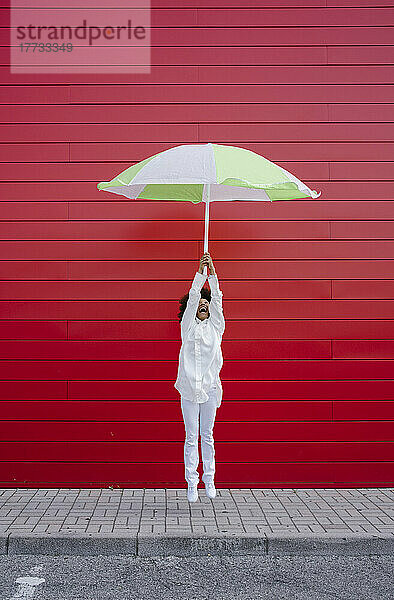 Woman jumping with umbrella on footpath in front of red wall