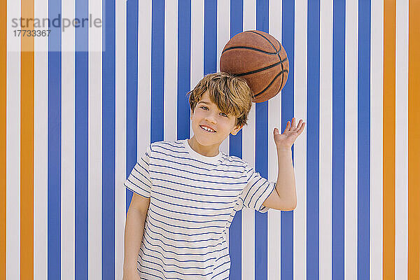 Verspielter Junge balanciert Basketball auf dem Kopf vor einer blau-orange gestreiften Wand