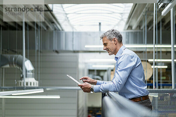 Geschäftsmann lehnt mit digitalem Tablet an Geländer in Fabrik