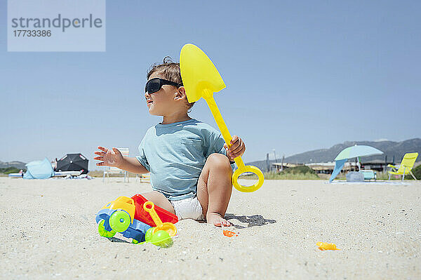 Junge mit Sonnenbrille und Schaufel am Strand an einem sonnigen Tag