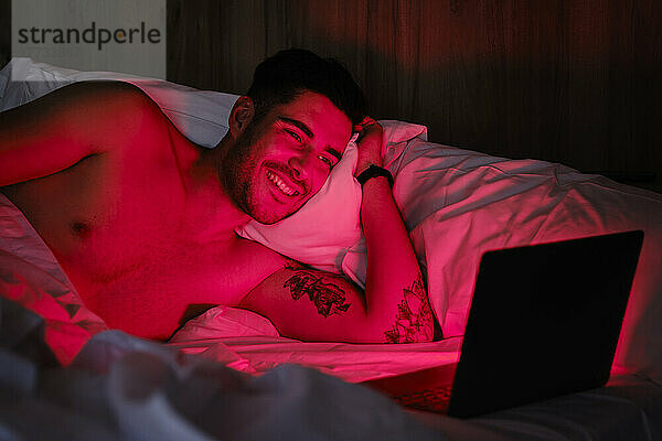 Lächelnder Mann blickt zu Hause im Bett auf einen rot beleuchteten Laptop