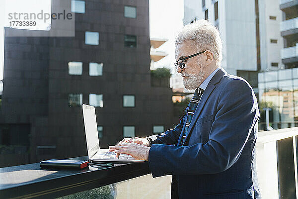 Leitender Geschäftsmann nutzt Laptop mit Solarbatterieladegerät und steht auf dem Bürobalkon