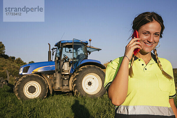 Lächelnde Bäuerin  die vor dem Traktor steht und mit dem Handy spricht