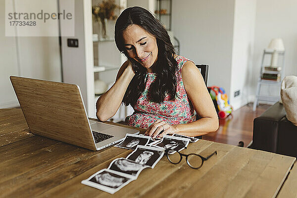 Lächelnde schwangere Frau sitzt mit Laptop und betrachtet Ultraschalluntersuchungen