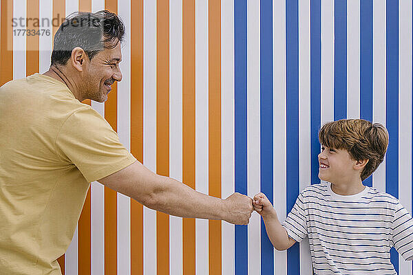 Lächelnder Mann gibt seinem Sohn vor einer orange-blau gestreiften Wand einen Fauststoß