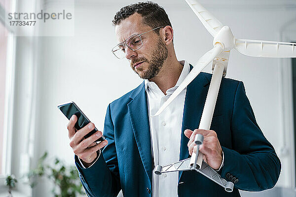 Geschäftsmann hält Windturbinenmodell mit Smartphone im Büro