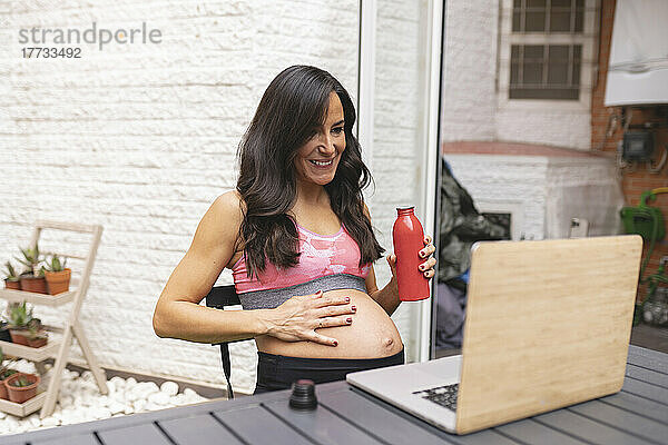Lächelnde schwangere Freiberuflerin mit Wasserflasche und Videoanruf über Laptop am Tisch auf dem Balkon