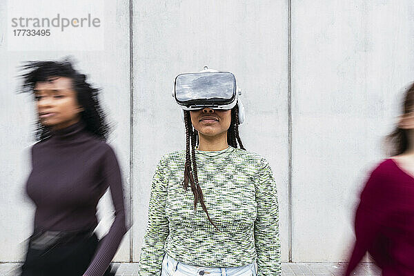 Smiling young woman wearing virtual reality simulator standing in front of wall