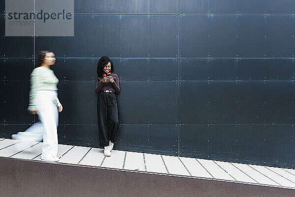 Glückliche Frau mit Afro-Frisur und Mobiltelefon  die vor der Wand steht