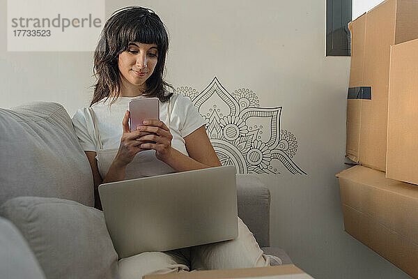 Frau sitzt mit Laptop auf Sofa im neuen Zuhause und benutzt Smartphone
