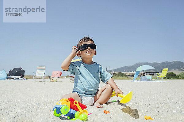 Junge passt Sonnenbrille an und sitzt mit Spielzeug im Sand am Strand