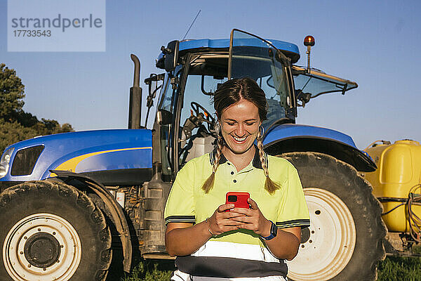 Glückliche Bäuerin  die an einem sonnigen Tag ihr Smartphone vor dem Traktor benutzt