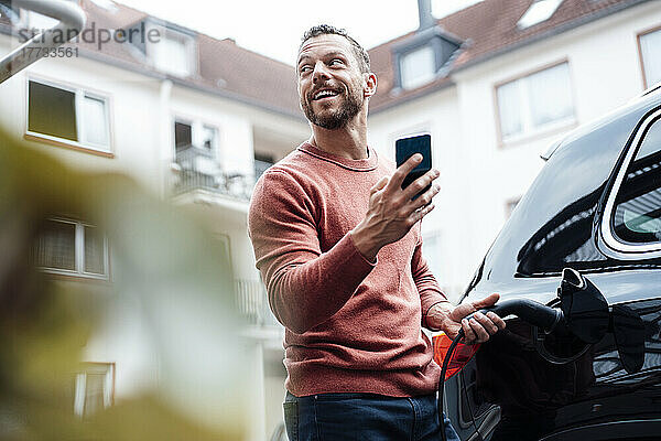 Glücklicher Mann mit Smartphone  der Elektroauto am Bahnhof auflädt