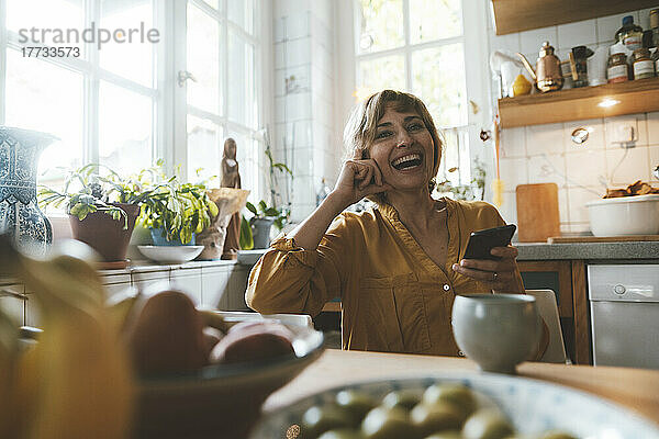 Glückliche reife Frau mit Smartphone sitzt am Esstisch