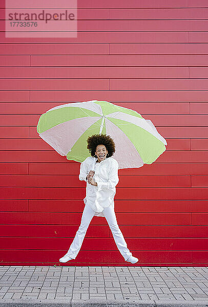 Fröhliche Frau mit Regenschirm springt vor rote Wand