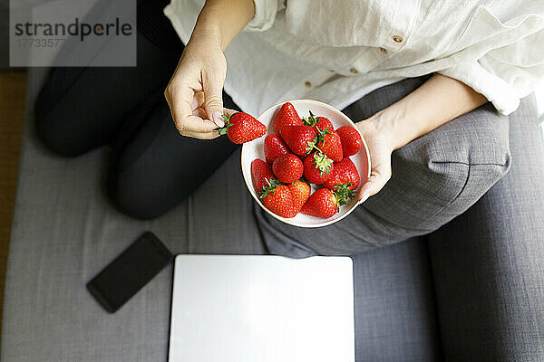 Frau hält eine Schüssel mit frischen Erdbeeren in der Hand und sitzt mit Laptop und Smartphone auf dem heimischen Sofa