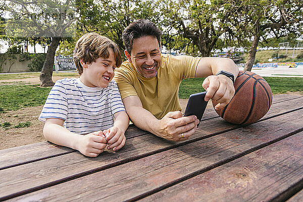 Lächelnder Mann mit Basketball zeigt seinem am Tisch sitzenden Sohn sein Mobiltelefon