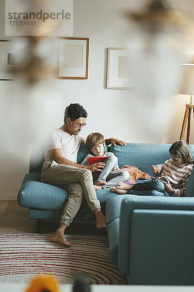 Vater sitzt mit Sohn und Tochter und nutzt Tablet-PC auf dem heimischen Sofa