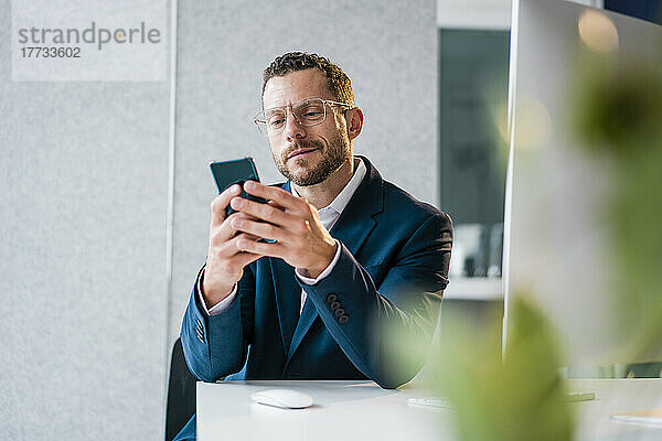 Lächelnder Geschäftsmann sitzt am Schreibtisch im Büro und nutzt sein Smartphone