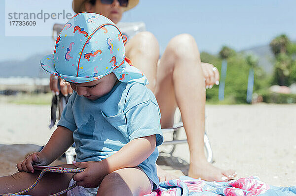Junge benutzt Mobiltelefon vor der Mutter und entspannt sich an einem sonnigen Tag am Strand