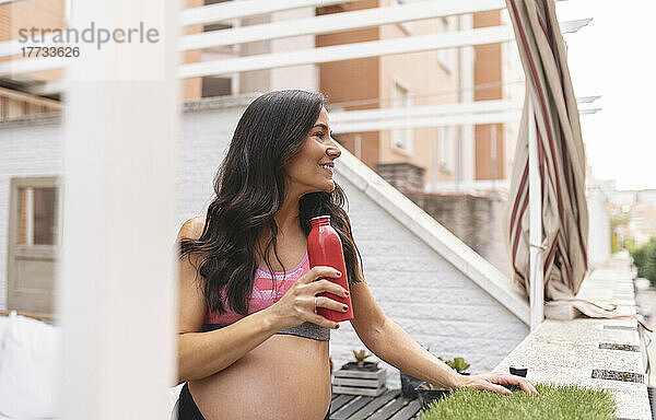 Lächelnde schwangere Frau mit Wasserflasche steht auf dem Balkon