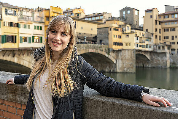 Lächelnde schöne Frau mit Pony steht an der Wand vor der Brücke Ponte Vecchio