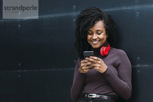 Smiling woman wearing wireless headphones using smart phone in front of wall