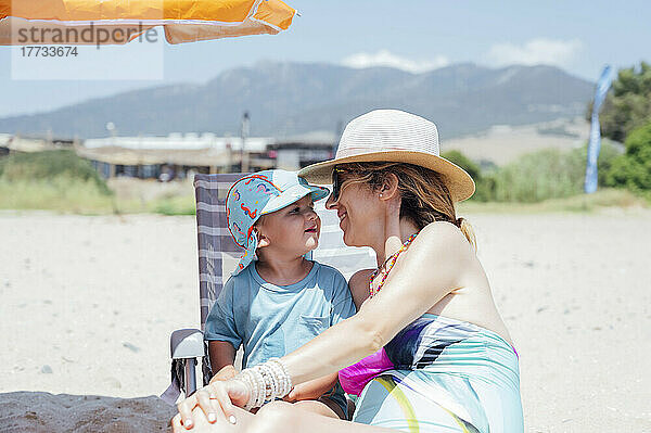 Glückliche Mutter mit Hut sitzt an einem sonnigen Tag mit ihrem Sohn am Strand