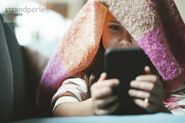 Mädchen benutzt Smartphone  das zu Hause unter einer Decke liegt