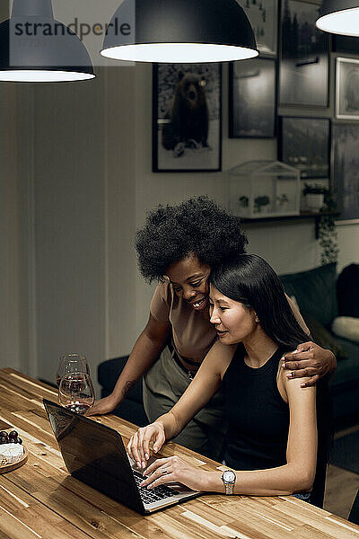 Lächelnde Frau umarmt Freundin mit Laptop am Tisch