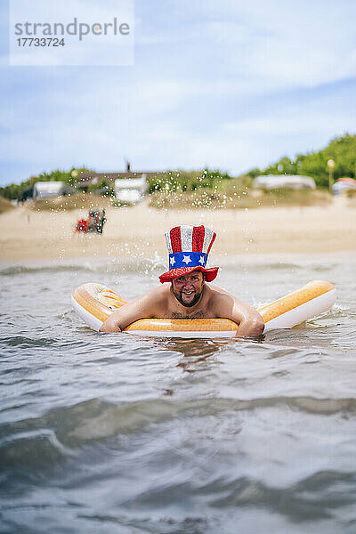 Lächelnder Mann mit Uncle Sam-Hut schwimmt mit Poolfloß im Meer