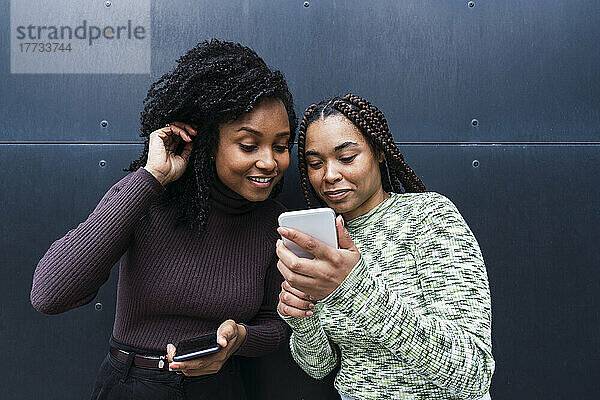 Lächelnde junge Frau mit Freundin  die ihr Smartphone vor der Wand benutzt